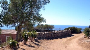 A rural setting with a dirt path leading to a stone wall, a small building with a tiled roof, and a view of the sea.