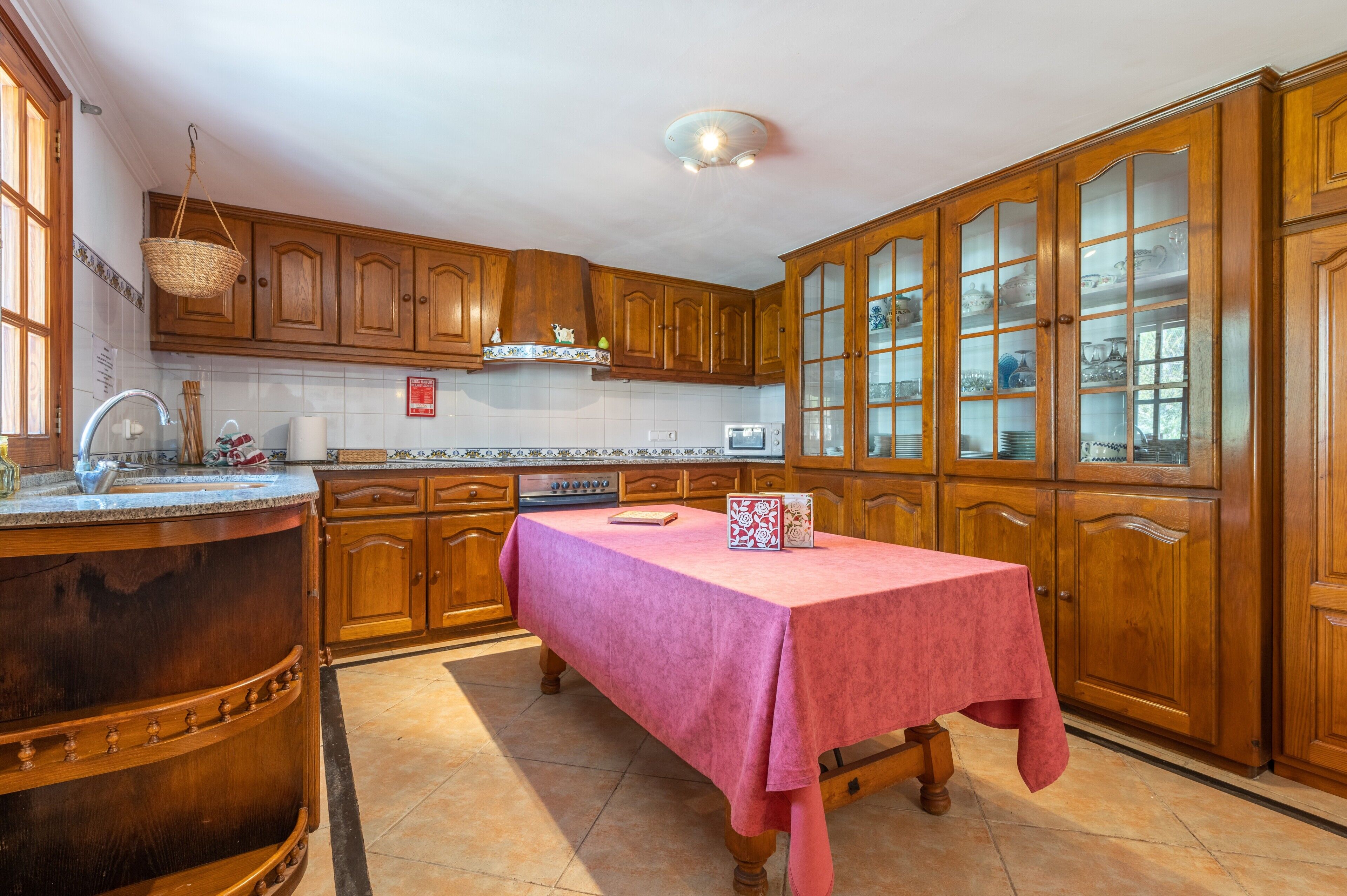 A kitchen with wooden cabinets, a marble countertop, a red tablecloth, and a hanging basket.