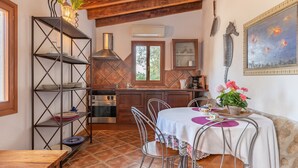 A rustic kitchen with a dining area, wooden beams, a metal shelf, and a painting. - Villa La Vinya Gran with Mountain View, Sauna, Pool, Jacuzzi, Wi-Fi, A/C, Terraces & Tennis Court (PUERTO DE POLLENSA)