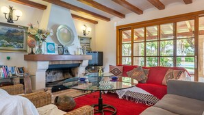 A living room with a fireplace, a glass coffee table, a red sofa, and a large window with a view of greenery. - Villa La Vinya Gran with Mountain View, Sauna, Pool, Jacuzzi, Wi-Fi, A/C, Terraces & Tennis Court (PUERTO DE POLLENSA)