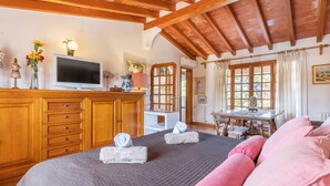 A bedroom with a bed, wooden cabinets, a television, a table, and a window with curtains. - Villa La Vinya Gran with Mountain View, Sauna, Pool, Jacuzzi, Wi-Fi, A/C, Terraces & Tennis Court (PUERTO DE POLLENSA)