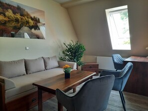 Uma sala de estar moderna com sofá, mesa de centro de madeira, escrivaninha e uma cadeira. Há uma grande janela que oferece luz natural e vista para árvores.