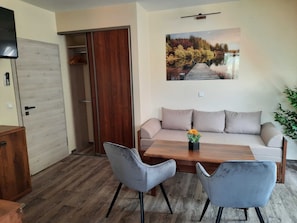 A room with a wooden floor, a beige sofa, two grey chairs, a wooden table with a vase of flowers, a wooden wardrobe, and a painting of a forest scene.