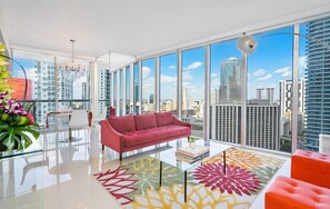 A modern living room with a red sofa, a glass coffee table, and a city view through large windows. - Brickell Open-Concept Suite for Groups (Miami)