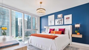 A modern bedroom with a large bed, a desk with a book, and a view of tall buildings. - Brickell Open-Concept Suite for Groups (Miami)