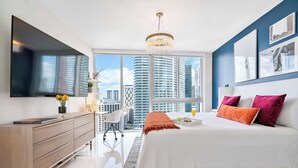 A modern bedroom with a large bed, a dresser, a TV mounted on the wall, and a city view through large windows. - Brickell Open-Concept Suite for Groups (Miami)