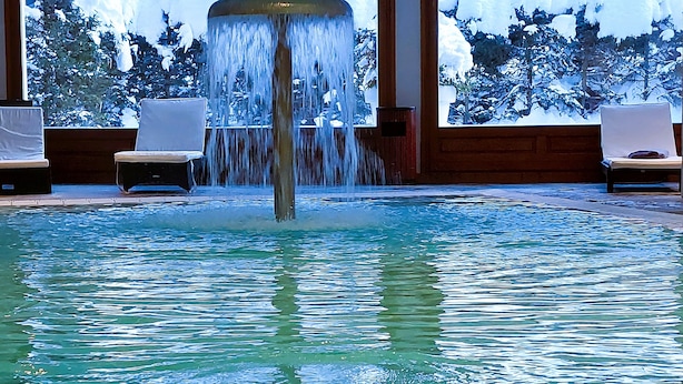 Una piscina con un elemento central de agua, rodeada de sillas de descanso y vistas a árboles nevados.
