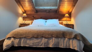 Un dormitorio con tragaluz, vigas de madera en el techo, dos mesitas de noche con lámparas, una cama con colcha estampada y almohadas blancas.