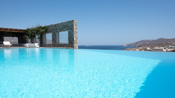 Una piscina infinita de color azul intenso con un arco de piedra y sillones de descanso.