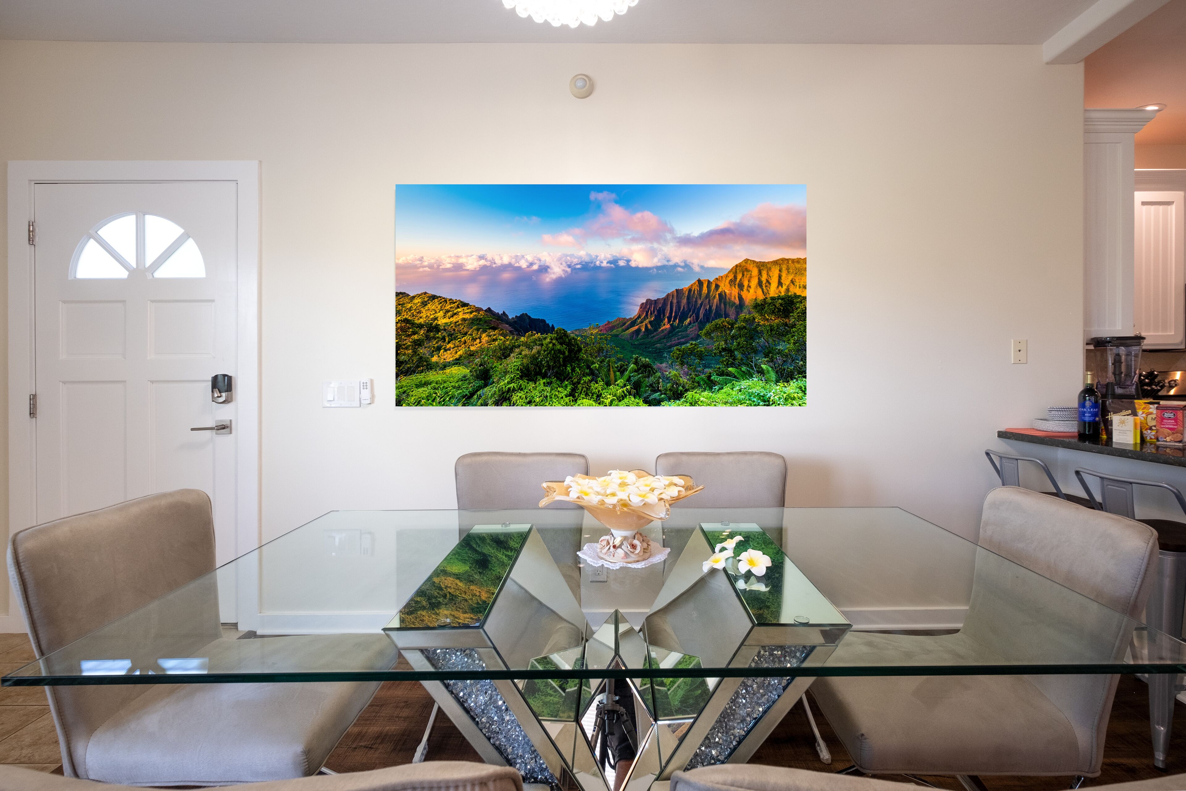 A dining area with a glass table, beige chairs, and a wall-mounted painting of a landscape.