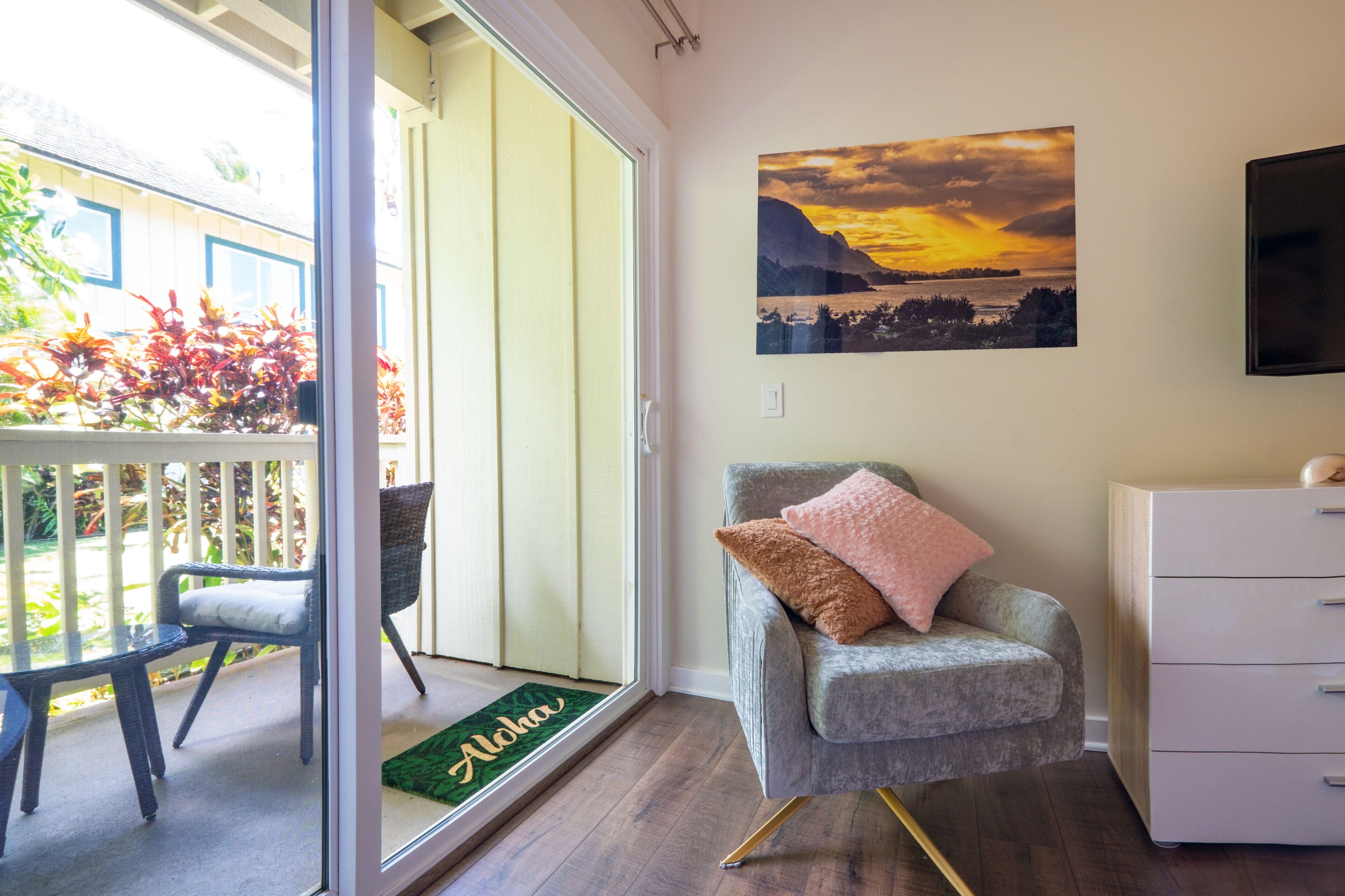 Et værelse med en skydedør, der f ører til en balkon, en grå lænestol med pude, en hvid kommode og et billede af en solnedgang i ramme på væggen.