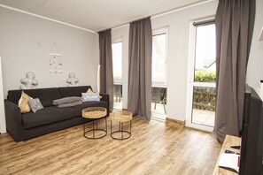 A living room with a sofa, two wicker side tables, a television, and a balcony with curtains.