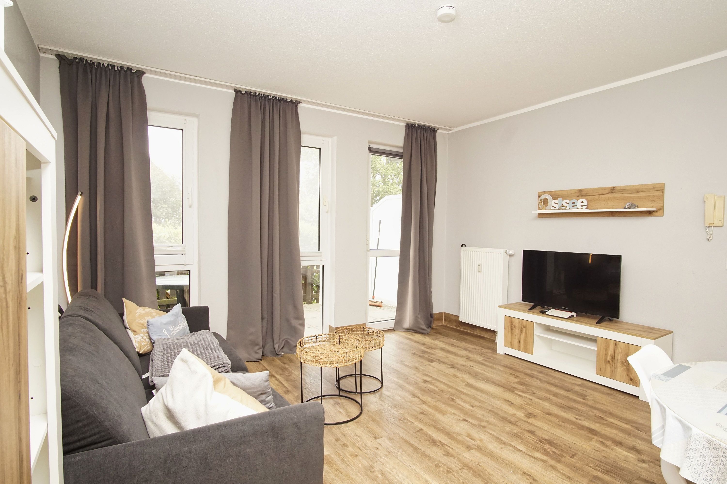 Un salon moderne comprenant un canapé, deux petites tables d’appoint en osier, un téléviseur posé sur un meuble en bois et de grandes fenêtres avec des rideaux.