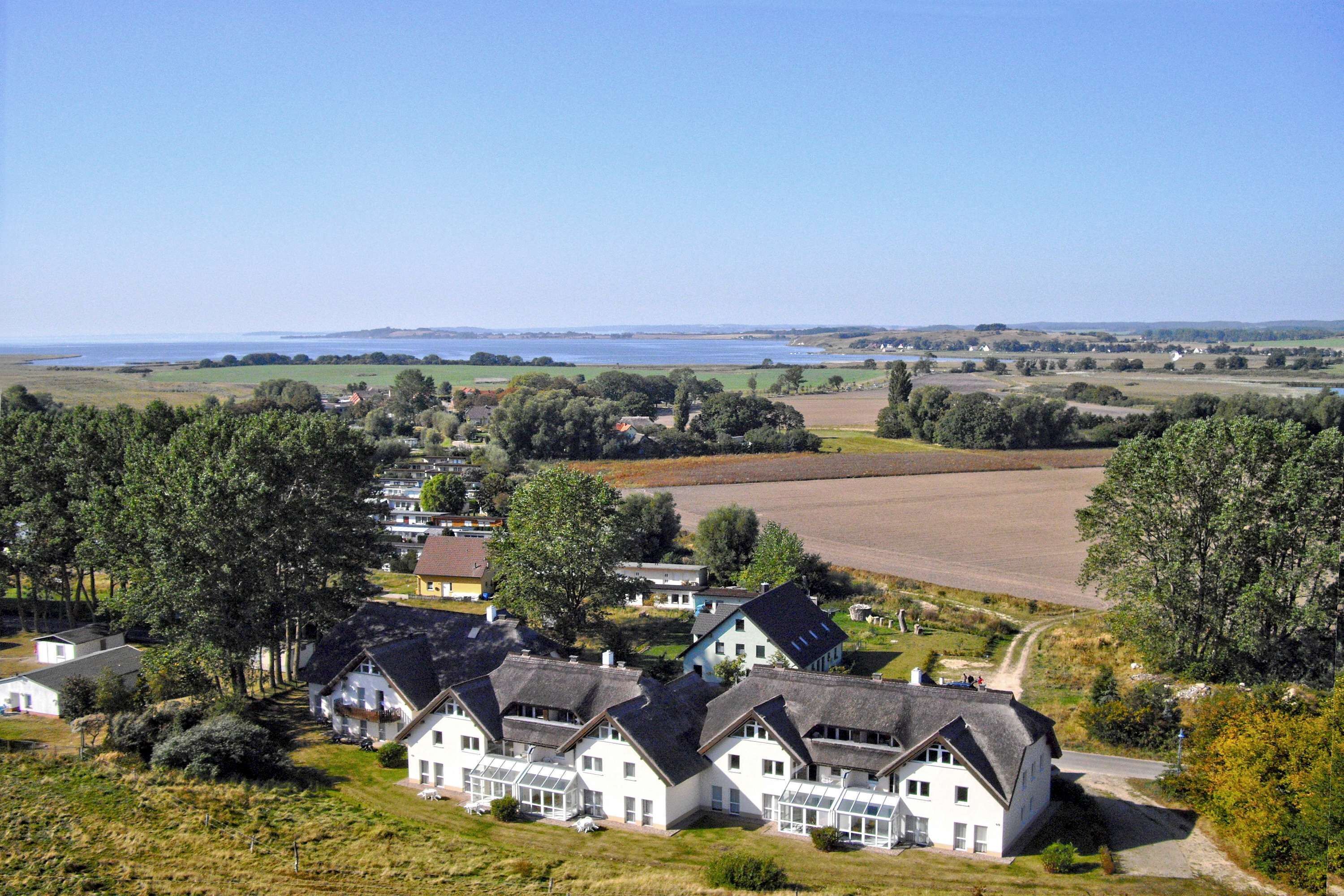 A cluster of white houses with dark roofs, surrounded by greenery and a view of a lake.