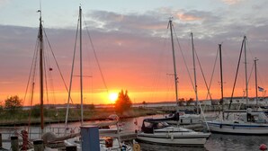Uma marina com vários barcos atracados, um pôr do sol ao fundo e uma área gramada com árvores.