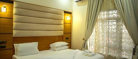 A hotel room with a large bed, wooden headboard, bedside lamps, an air conditioning unit, and patterned curtains.