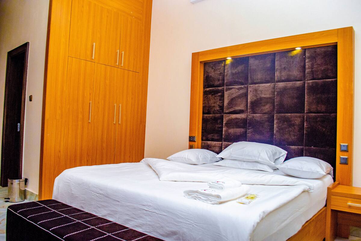 Uma cama bem arrumada com lençóis brancos, cabeceira de madeira e mesa de cabeceira com abajur.
