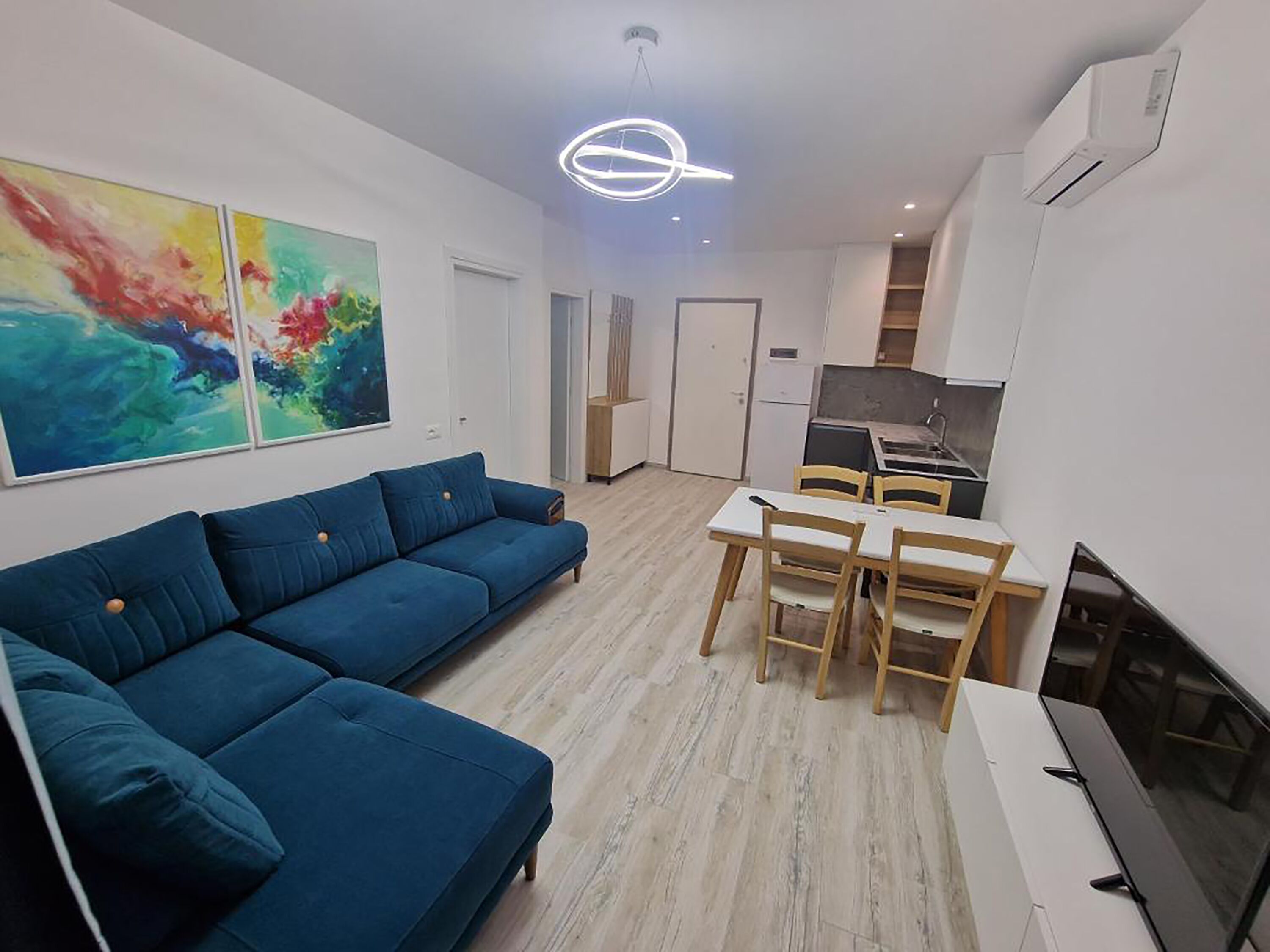 A modern living space with a blue sofa, a dining table with chairs, a television, and a kitchen area.