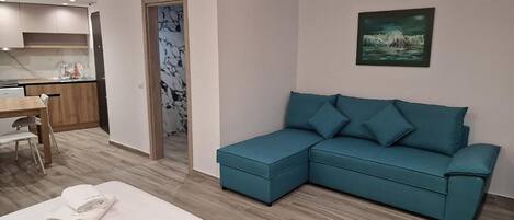 A modern hotel room with a large teal sectional sofa, a neatly made bed with white linens, a dining area with a wooden table and chairs, and a kitchen area in the background.