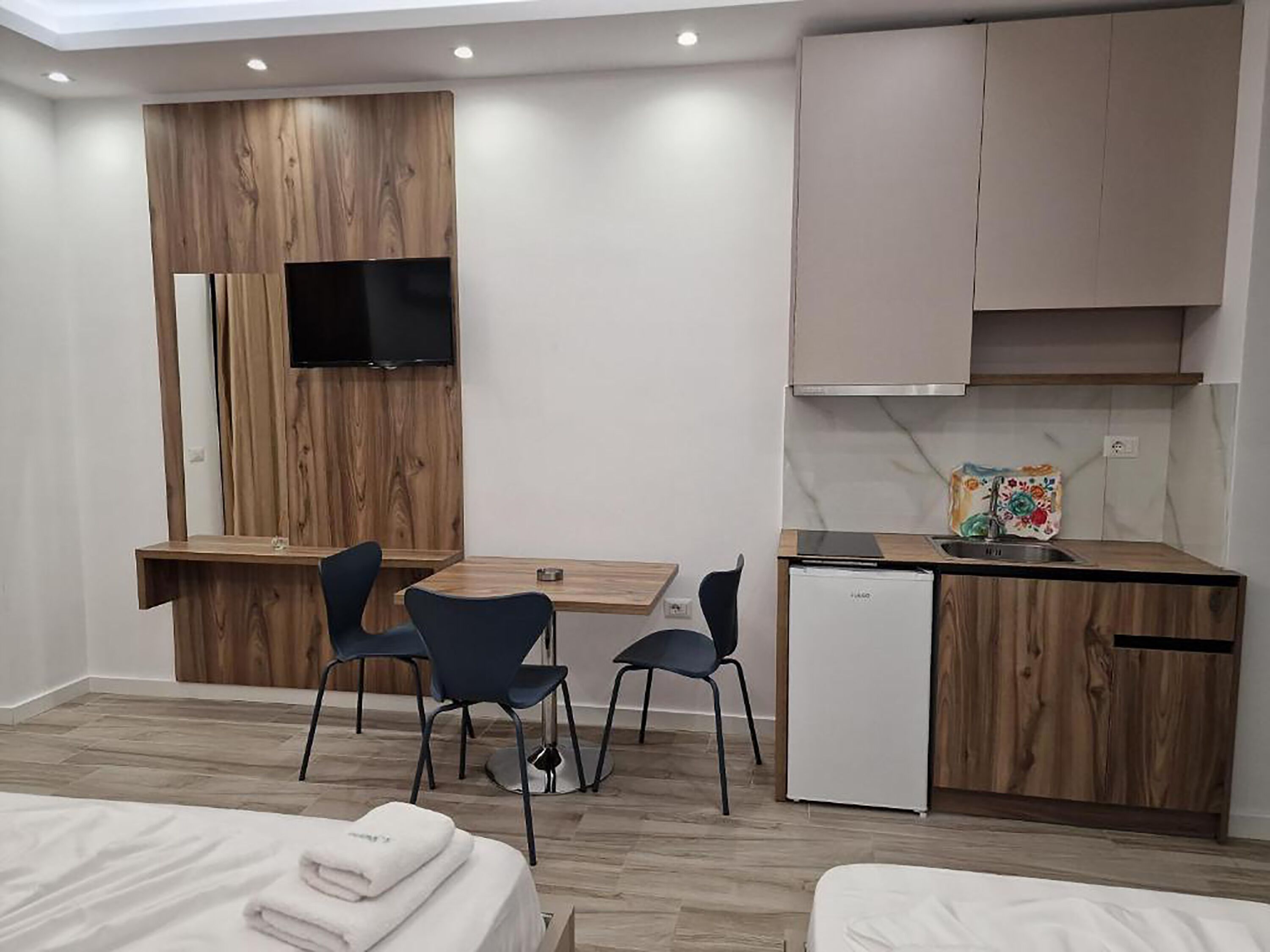 A modern hotel room with a wooden desk, two chairs, a small kitchenette, and a bed with neatly folded towels.