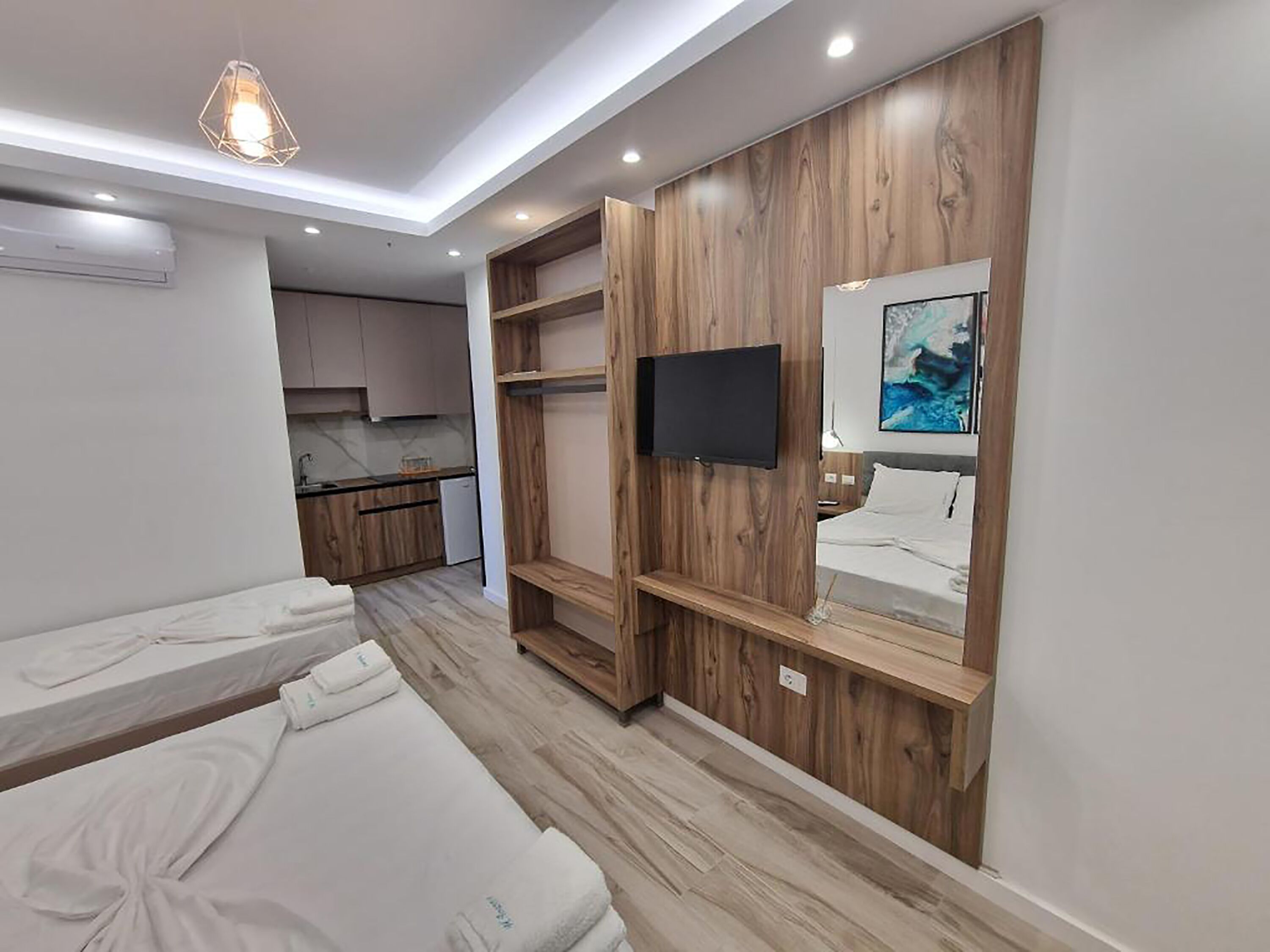 A modern hotel room with a kitchenette, a TV mounted on a wooden panel, and two beds.