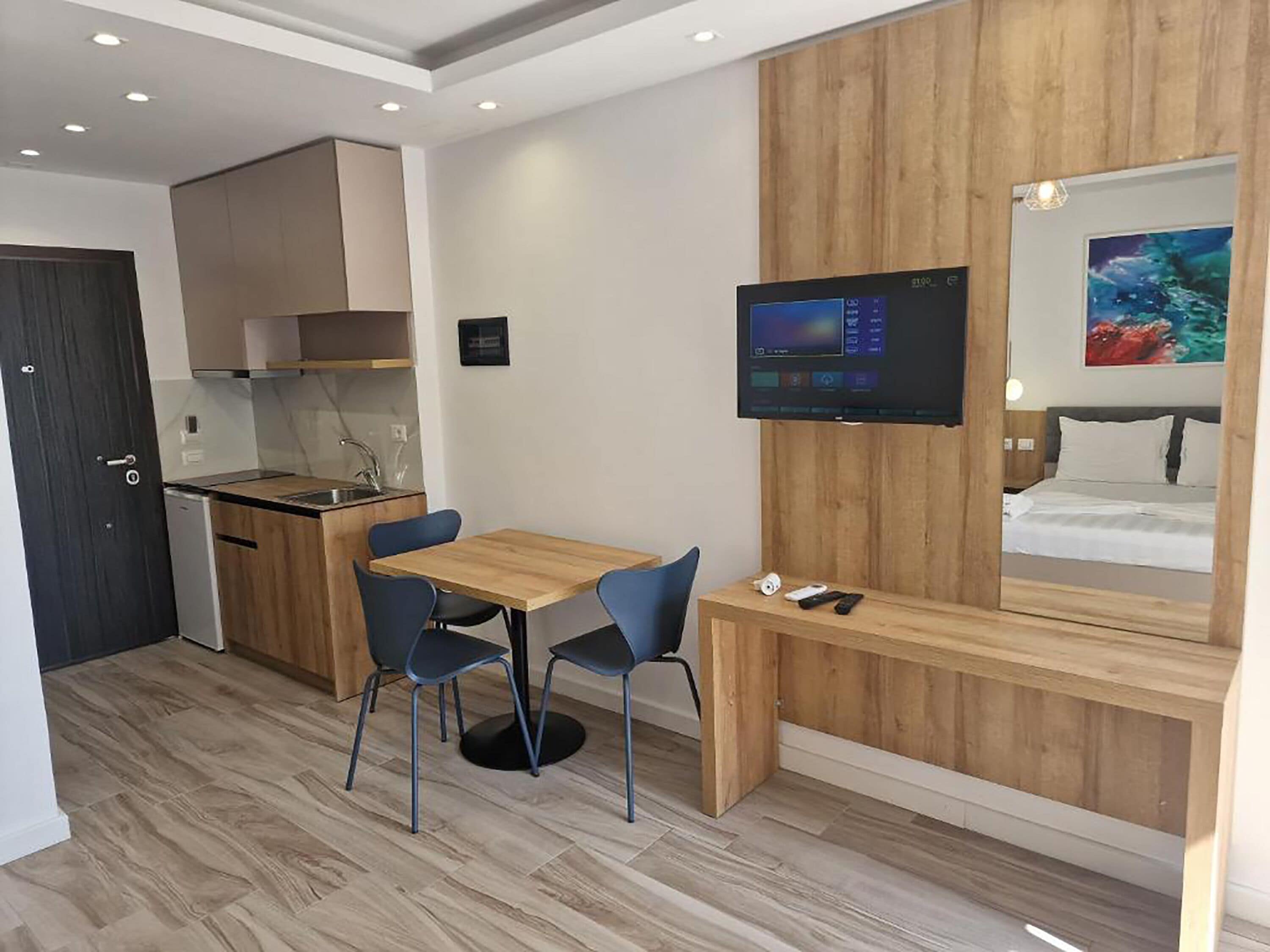 Ein modernes Hotelzimmer mit einem Holztisch, zwei Stühlen, einem an der Wand montierten Fernseher, einem Bett mit Kopfteil und einer kleinen Küchenzeile.