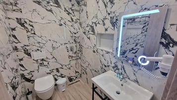 Un baño con pared de mármol, inodoro blanco, lavabo con espejo y zona de ducha.