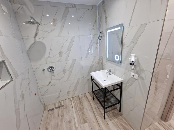 A modern bathroom with a marble shower, a white sink on a black metal stand, and a mirror with a light. - Joanna Suites (Sarandë)
