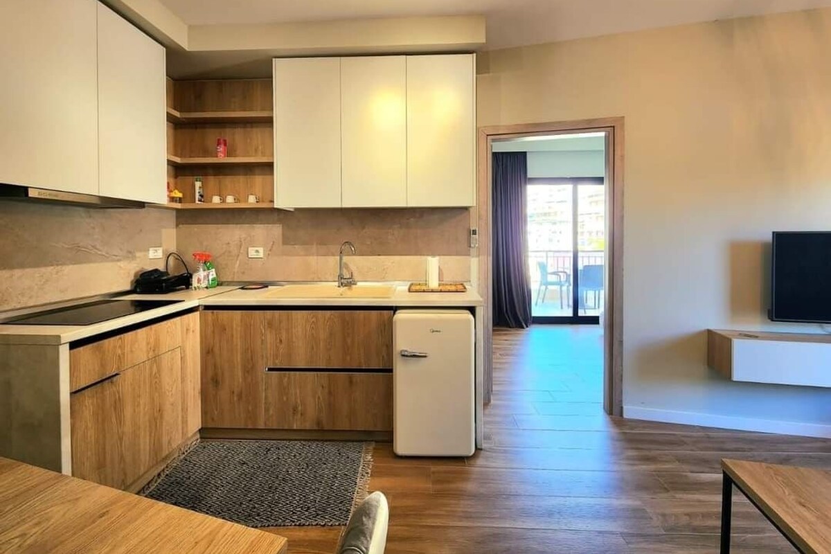 A modern kitchen with wooden cabinets, a built-in oven, and a small refrigerator. There is a dining area with a table and chairs, and a TV mounted on the wall.