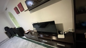 Ett hotellrum med en säng, en tv, ett skrivbord med en dator, en hylla med dekorativa föremål och en väggmonterad lampa.