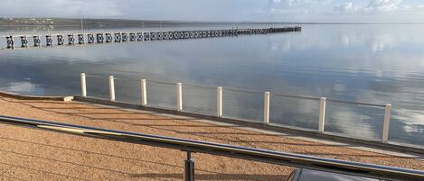 A coastal view with a pebbled beach, a metal railing, and a pier extending into calm waters.