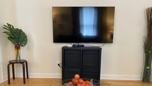 A living room with a flat-screen TV mounted on the wall, a glass coffee table with oranges, a dark cabinet, and a blue patterned rug. - Calm Relaxing Home Away from Home   (Westfield)