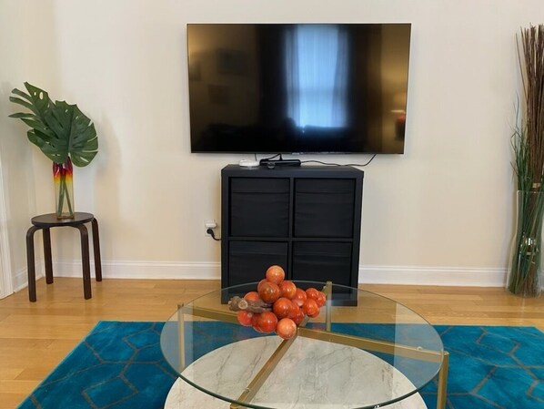 Una sala de estar con un televisor de pantalla plana montado en la pared, una mesa de centro de vidrio con naranjas, un mueble oscuro y un tapete azul estampado.