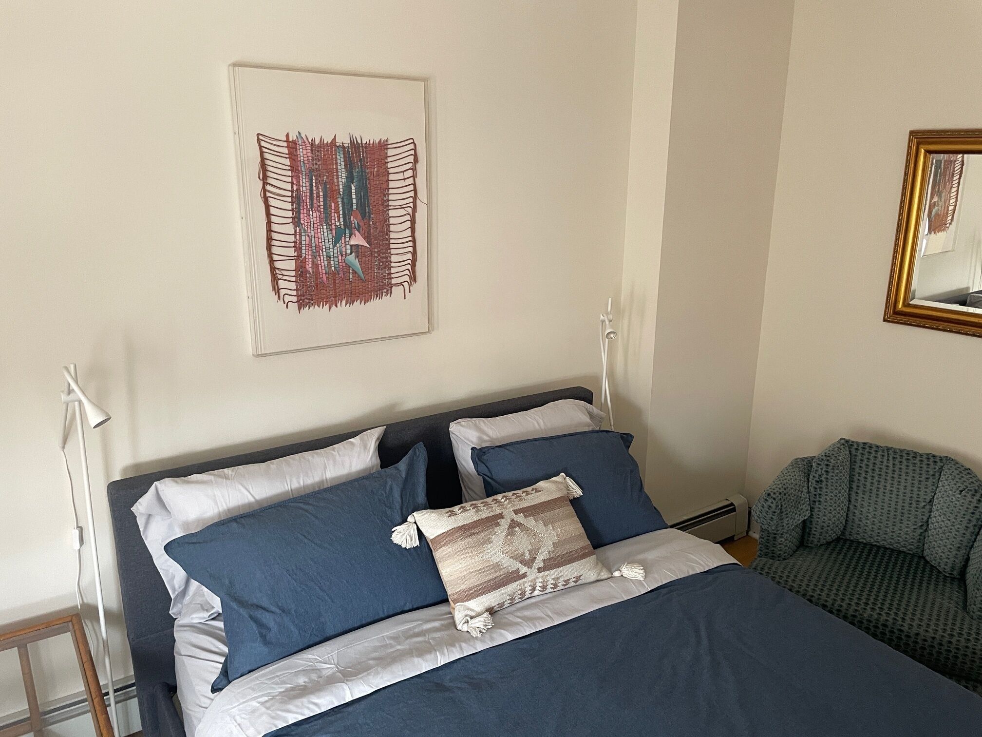 A neatly made bed with blue and white bedding, a patterned throw pillow, a framed abstract artwork on the wall, and a mirror with a gold frame.