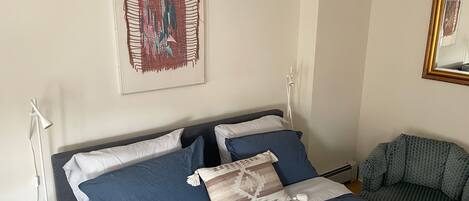 A neatly made bed with blue and white bedding, a patterned throw pillow, a framed abstract artwork on the wall, and a mirror with a gold frame.