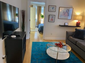 Una sala de estar moderna con un televisor de pantalla plana, una mesa de centro de vidrio con flores rojas, un sofá gris y una alfombra azul estampada.