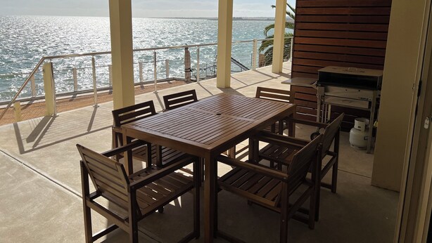 A covered outdoor area with a table and chairs, a grill, and a view of the sea.