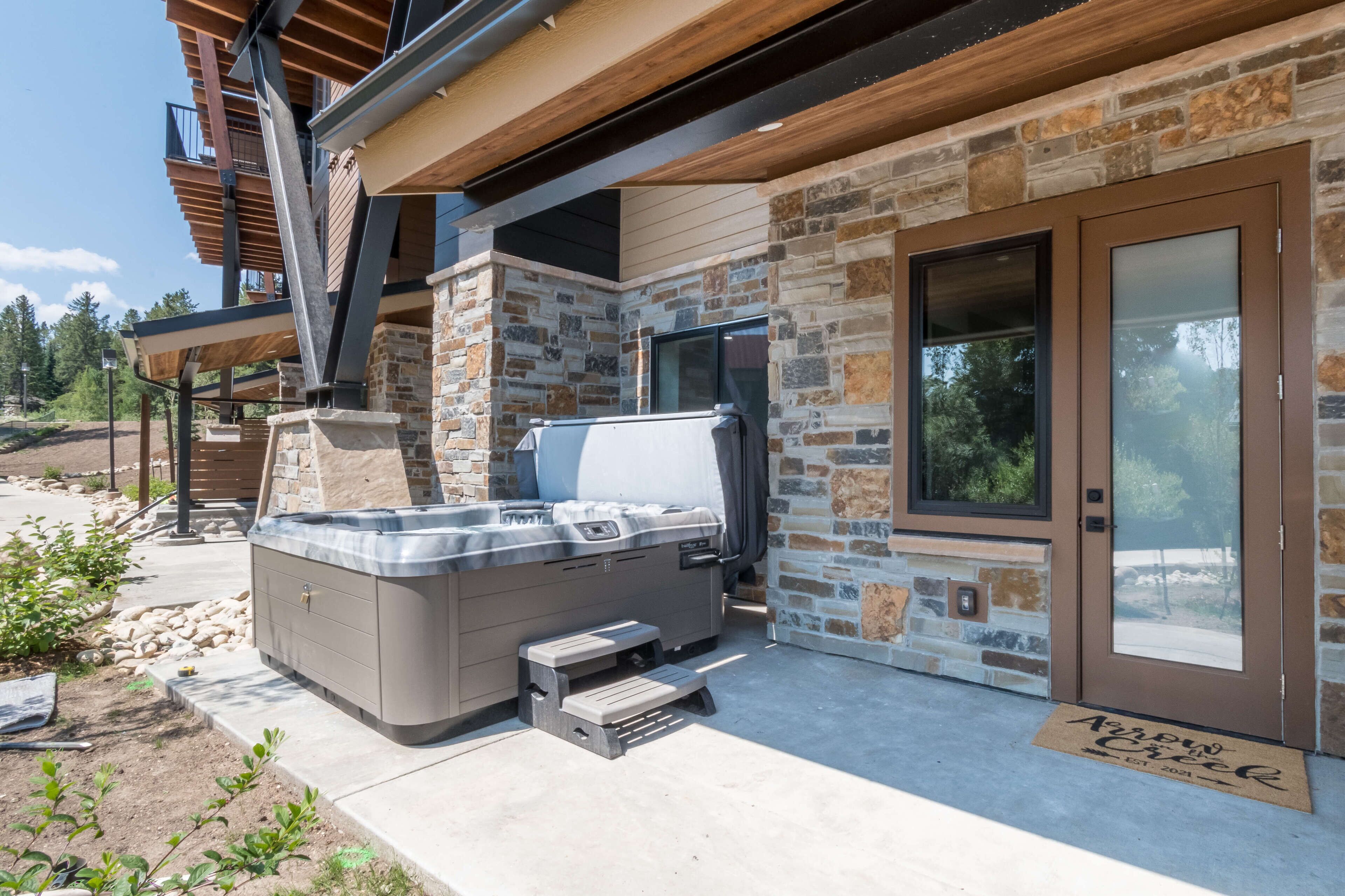 A modern outdoor hot tub area with a stone wall backdrop, a wooden deck, and a sliding glass door leading to a covered patio.