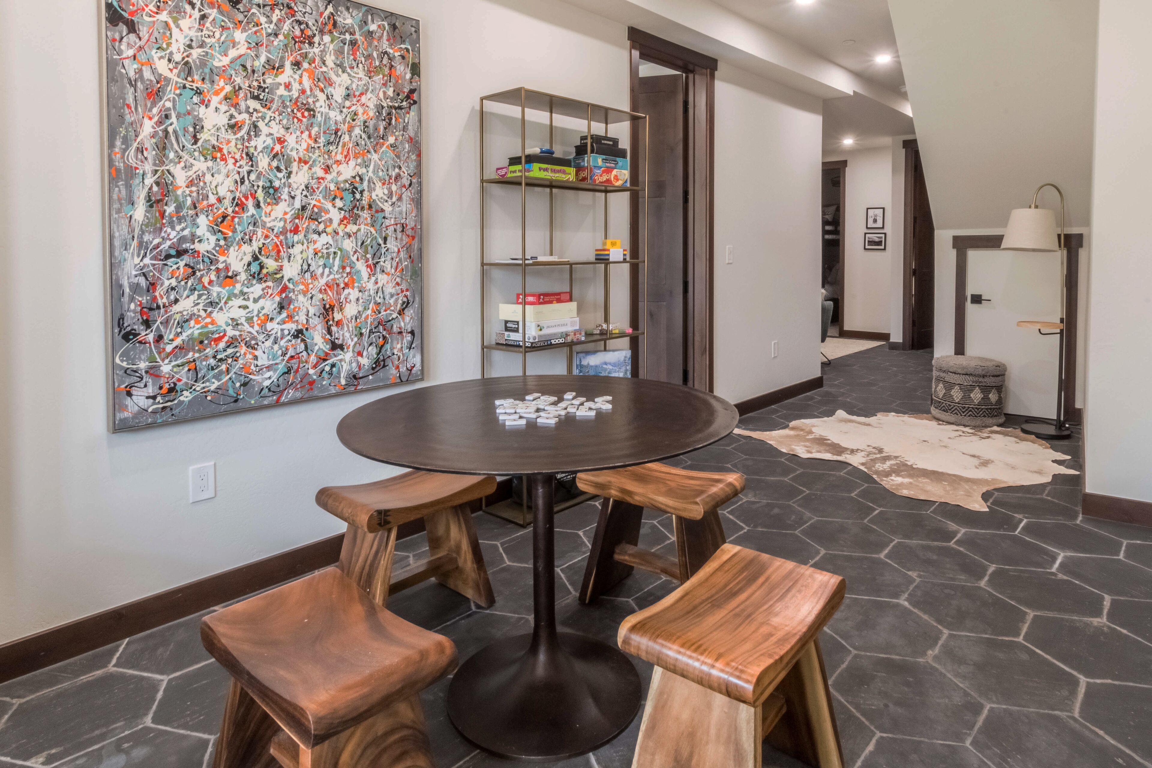 A modern dining area with a round table and wooden stools, a large abstract painting on the wall, and a bookshelf with various items.