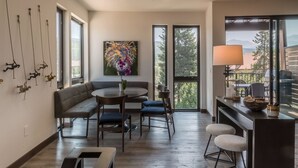A modern dining area with a round table, blue chairs, and a bench. There is a colorful abstract painting on the wall and a window with a view of trees and mountains.