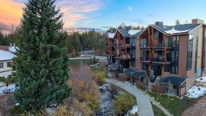 A modern multi-story building with balconies and large windows, surrounded by trees and a small stream.