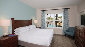 Una habitación de hotel con una cama grande, dos mesitas de noche con lámparas, un televisor, una silla y una ventana con vistas a una piscina y palmeras.
