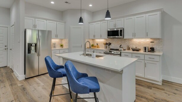 A modern kitchen with white cabinets, a stainless steel refrigerator, a microwave, and a large island with blue bar stools. - Modern and Comfortable Home Close to Downtown (Dallas)