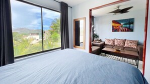 A bedroom with a large bed, a sofa, a painting, and a view of greenery and buildings.