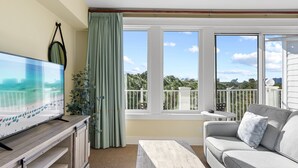 Un balcón con vistas a un paisaje urbano, un sofá gris, un televisor sobre un mueble de madera y una mesa con una planta.