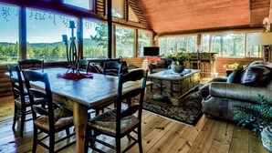 A cozy cabin interior with a wooden dining table, chairs, a sofa, and a TV. Large windows offer a view of the surrounding forest. - TrailsEndRanch ⛰Views, HotTub, Horseback riding on 1000's ac (Angel Fire)
