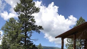 A cabin with a wooden deck, surrounded by a grassy field and trees, with mountains in the background. - TrailsEndRanch ⛰Views, HotTub, Horseback riding on 1000's ac (Angel Fire)