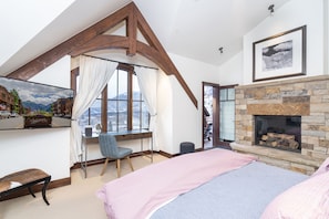 Une chambre avec une cheminée en pierre, un bureau avec une chaise, un lit et une grande fenêtre avec des rideaux.