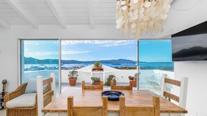 A dining area with a wooden table and chairs, potted plants, and a view of the sea and mountains through large windows.