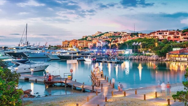 A marina with boats docked, a wooden pier, and buildings in the background.